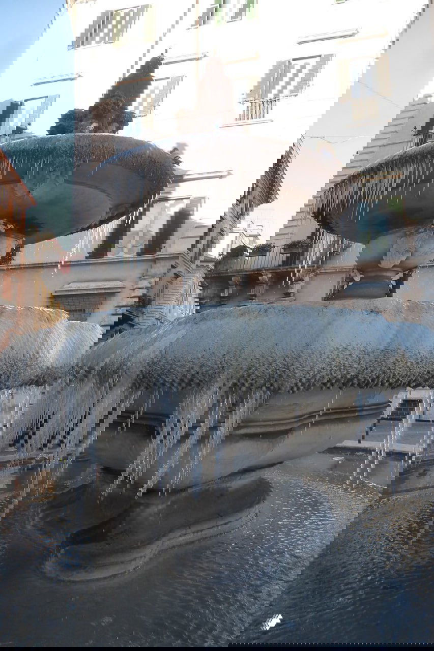 Фотография замёрзшего фонтана в Риме с сосульками и инеем на чашах фонтана, на фоне исторических зданий площади.