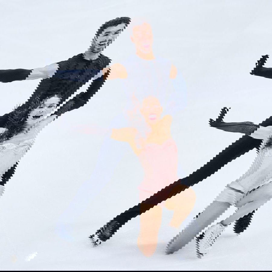 Парная постановка на льду: женщина в светло-розовом корсетном костюме опущена на колено, партнёр в чёрном позади, кадр передаёт динамику и взаимодействие партнёров.