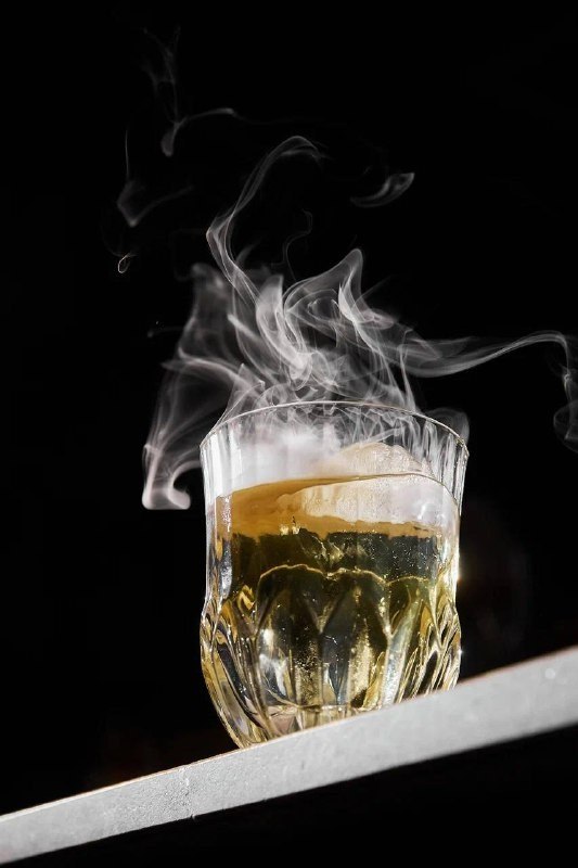 Коктейль в витом стеклянном бокале с лимонной долькой и клубами дыма на тёмном фоне, элегантная подача и акцент на аромате напитка.