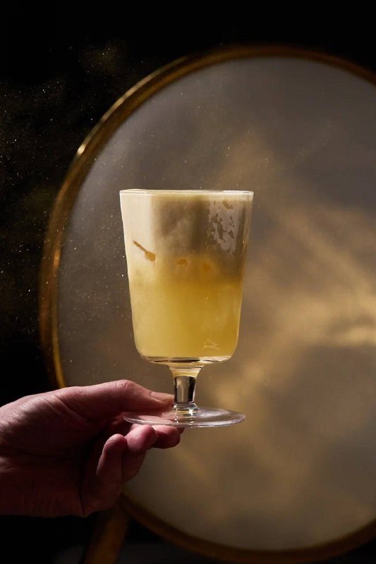 Рука держит высокий бокал с кремово-жёлтым коктейлем и лёгкой пеной; мягкий свет и круглая отражающая поверхность подчёркивают текстуру напитка.