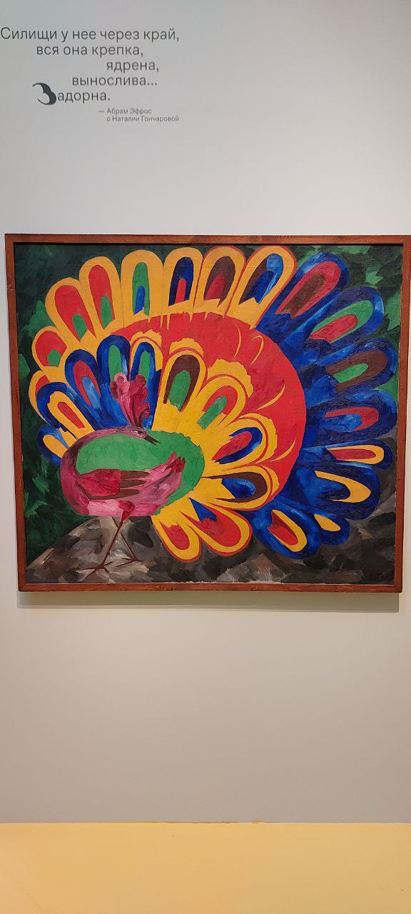 Яркая картина с изображением павлина и разноцветными перьями в насыщенных жёлто-красно-синих тонах, снятая в зале выставки.