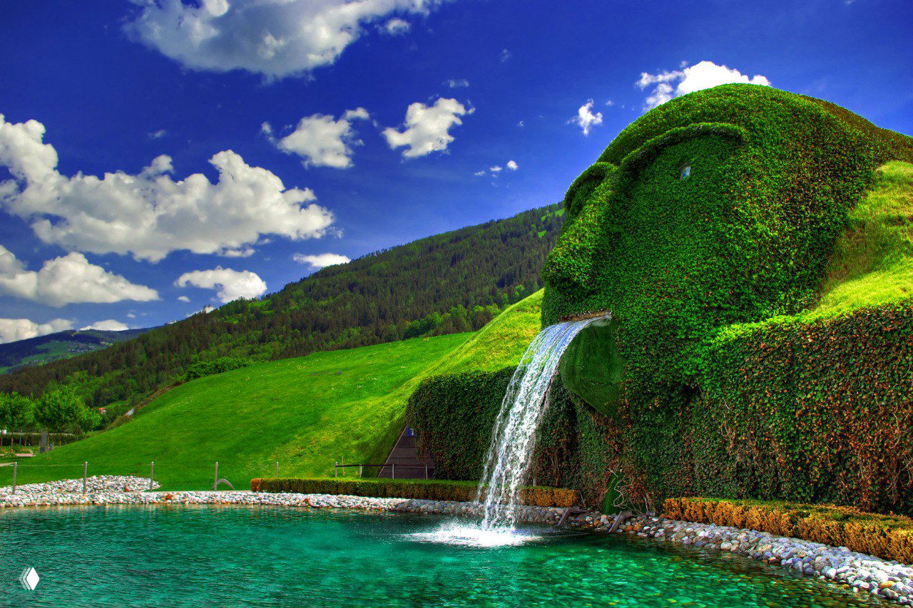 Крупный ракурс зелёной скульптуры‑гиганта музея Swarovski, из её рта бьёт водный каскад в бирюзовый пруд среди альпийского ландшафта.