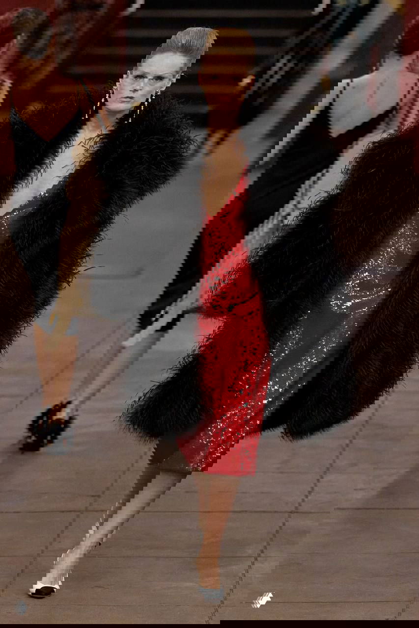 Драматичный образ: пышное чёрное пальто и красное блестящее платье на модели, проходящей по платформе метро; кадр с показа Métiers d’art Chanel.