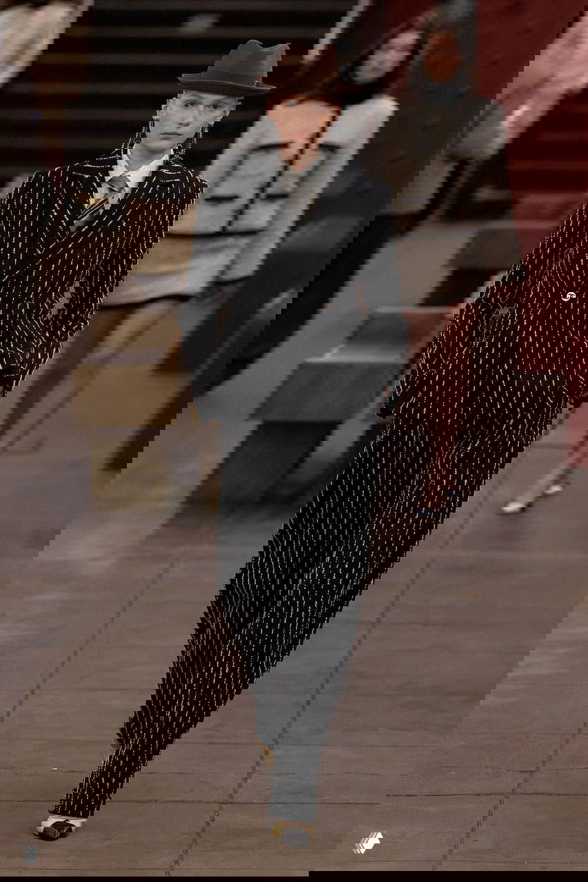 Модель в полосатом двубортном костюме и фетровой шляпе идёт по платформе метро; образ сочетает мужской крой и женскую элегантность, фото с показа.