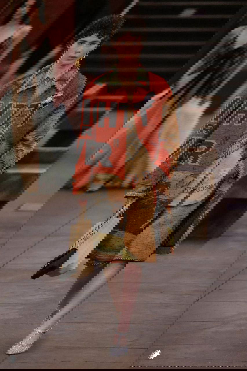 Модель в ярком пальто с принтом и туфлях идет по подиуму, имитирующему станцию метро, эпатажные детали и графичный принт заметны на образе.