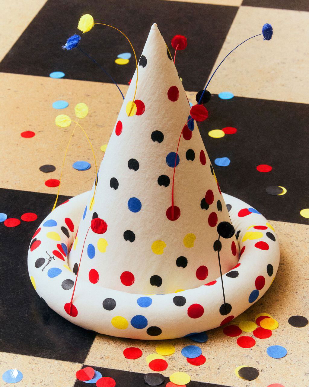 Крупный план белой шляпы‑конуса с разноцветными круглыми пятнами и бумажным конфетти на чёрно‑белом шахматном полу — деталь показа и оформления.