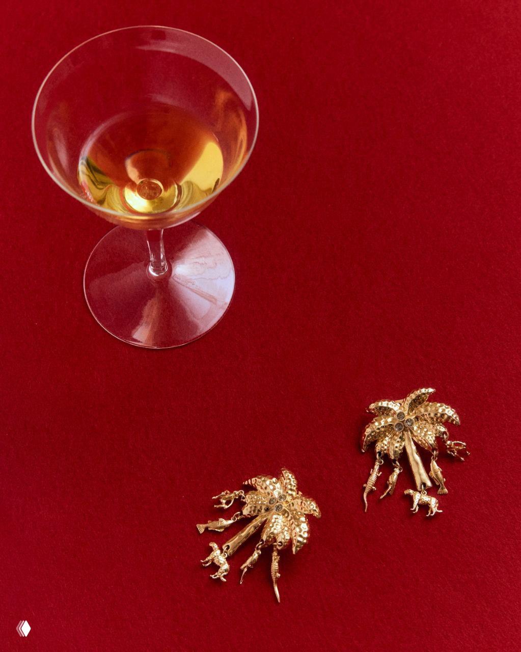 Коктейльный бокал с янтарной жидкостью и пара золотистых брошей на красной бархатной ткани — стилизованная натюрмортная деталь показа.