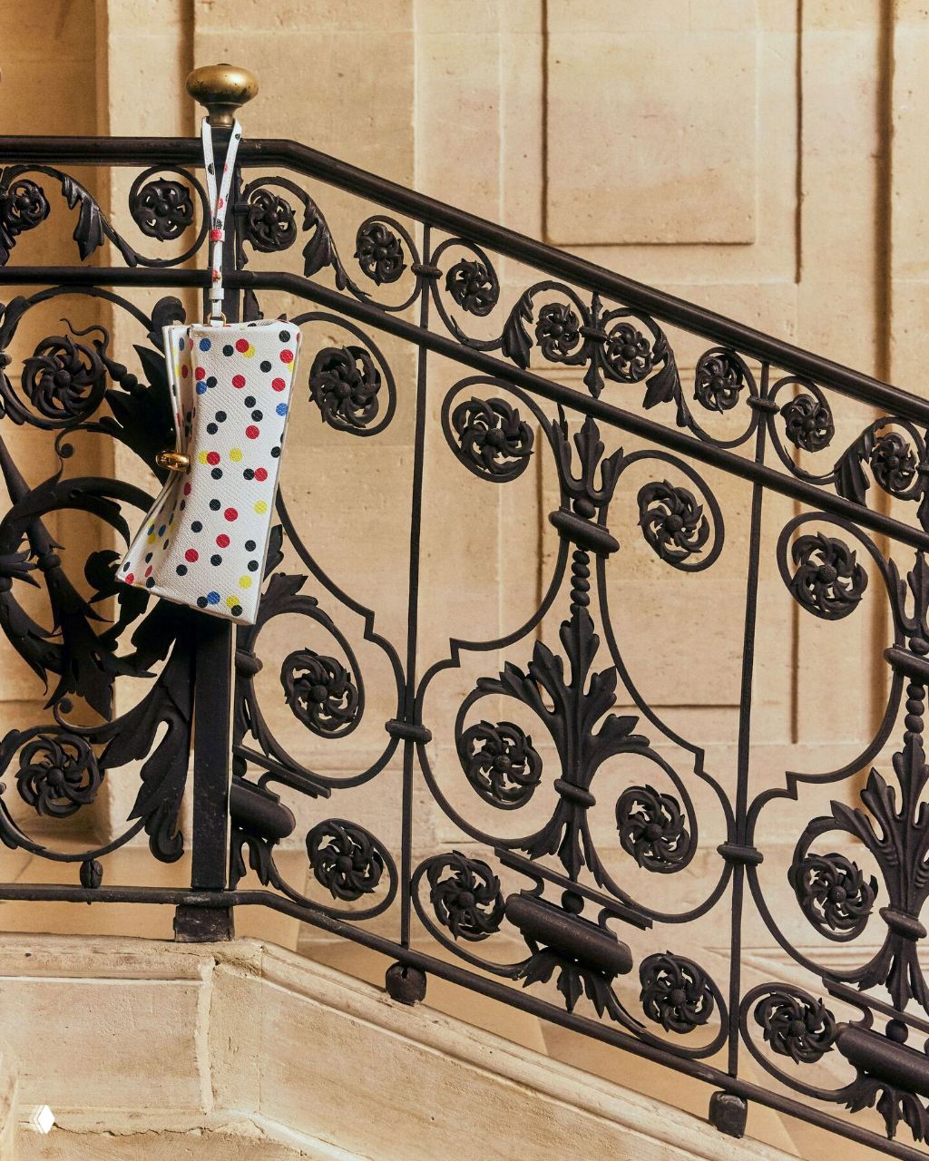 Миниатюрная сумка с разноцветным гороховым принтом, подвешенная на кованой чёрной ограде лестницы музея; сочетание дизайна и классической архитектуры.
