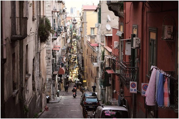Узкая неаполитанская улица с развешенным бельём, припаркованными машинами и плотной застройкой — типичный городской пейзаж.