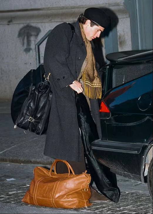 Мужчина в длинном тёмном пальто с большой дорожной сумкой рядом с автомобилем, кадр показывает дорожный стиль и аксессуары для поездок.