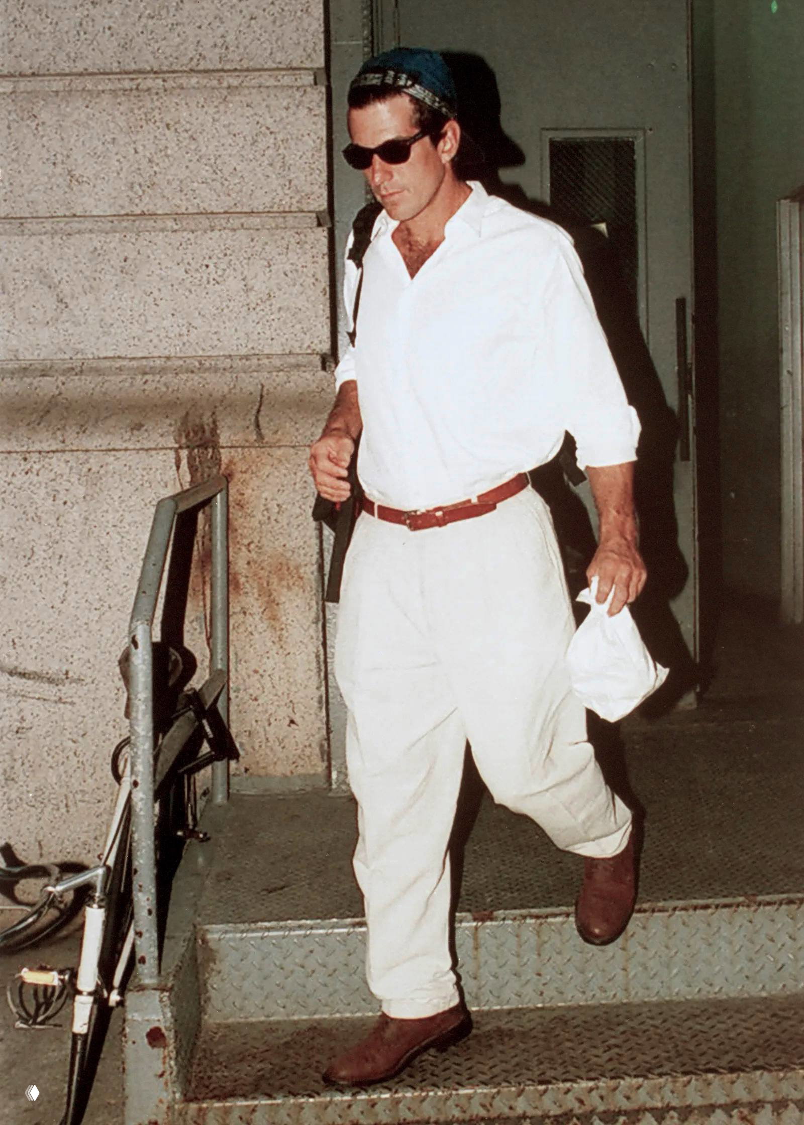 Мужчина в светлой рубашке и свободных брюках со слегка заправленным ремнём спускается по ступенькам — спокойный повседневный летний образ.