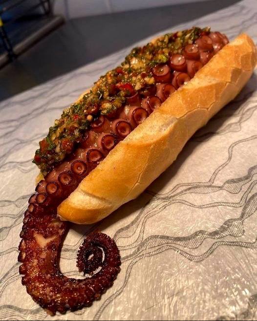Булка с приготовленным щупальцем осьминога и соусом сверху — смелое гастрономическое сочетание, акцент на текстуре и необычном блюде на столе.