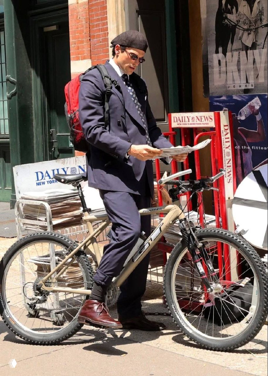 Мужчина в двубортном пиджаке и кепке, с рюкзаком и газетой у стойки с прессой, образ городской повседневной одежды с элементами формального костюма.