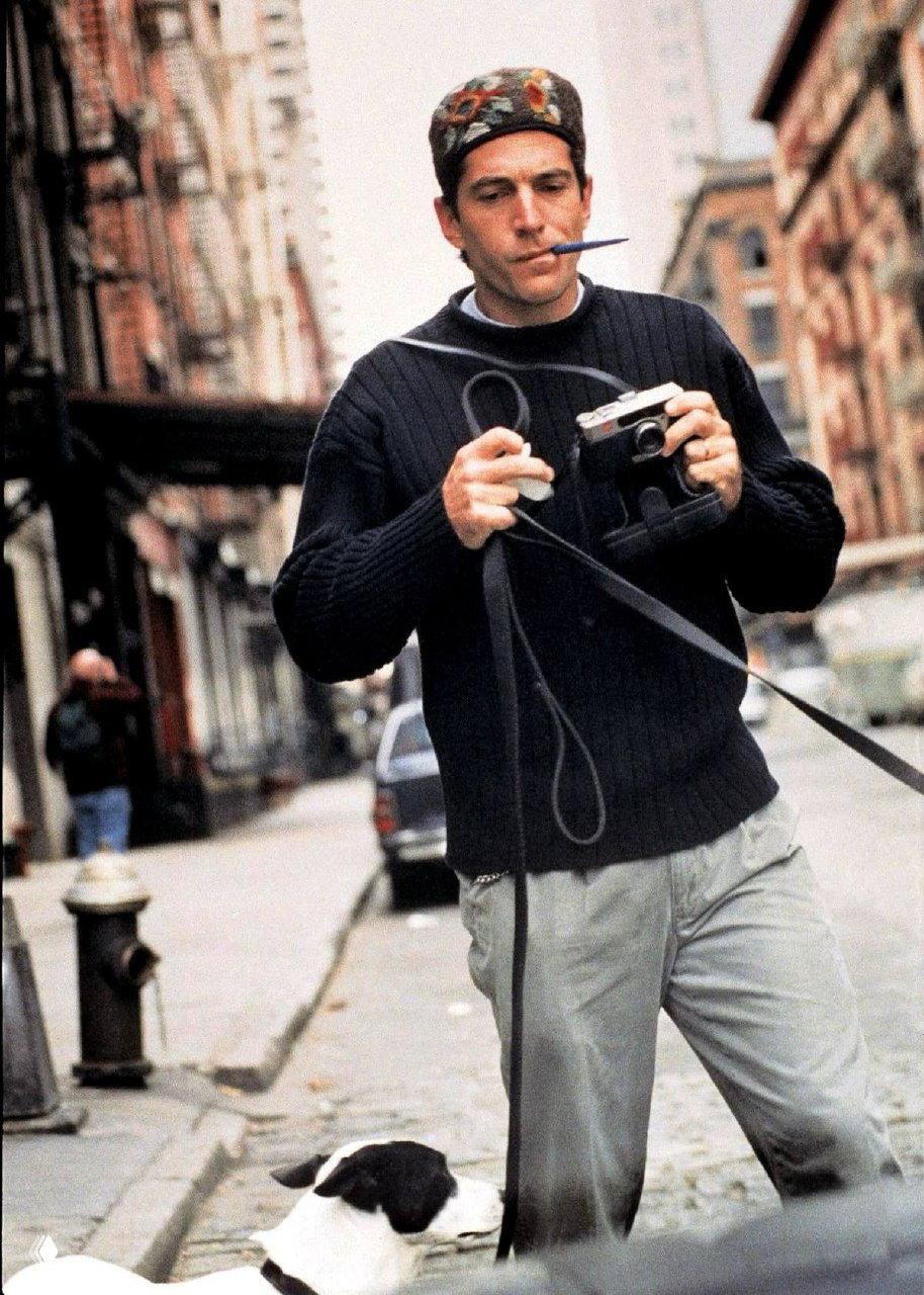 Уличный кадр: мужчина в тёмном свитере с фотоаппаратом и шнуром ремня, вокруг городская улица и собака у ног — типичный повседневный стиль.