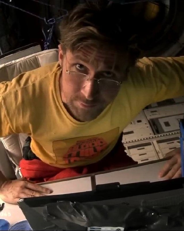 Крупный план главного героя в салоне космического корабля: мужчина в жёлтой футболке смотрит в камеру, вокруг элементы оборудования и панели.