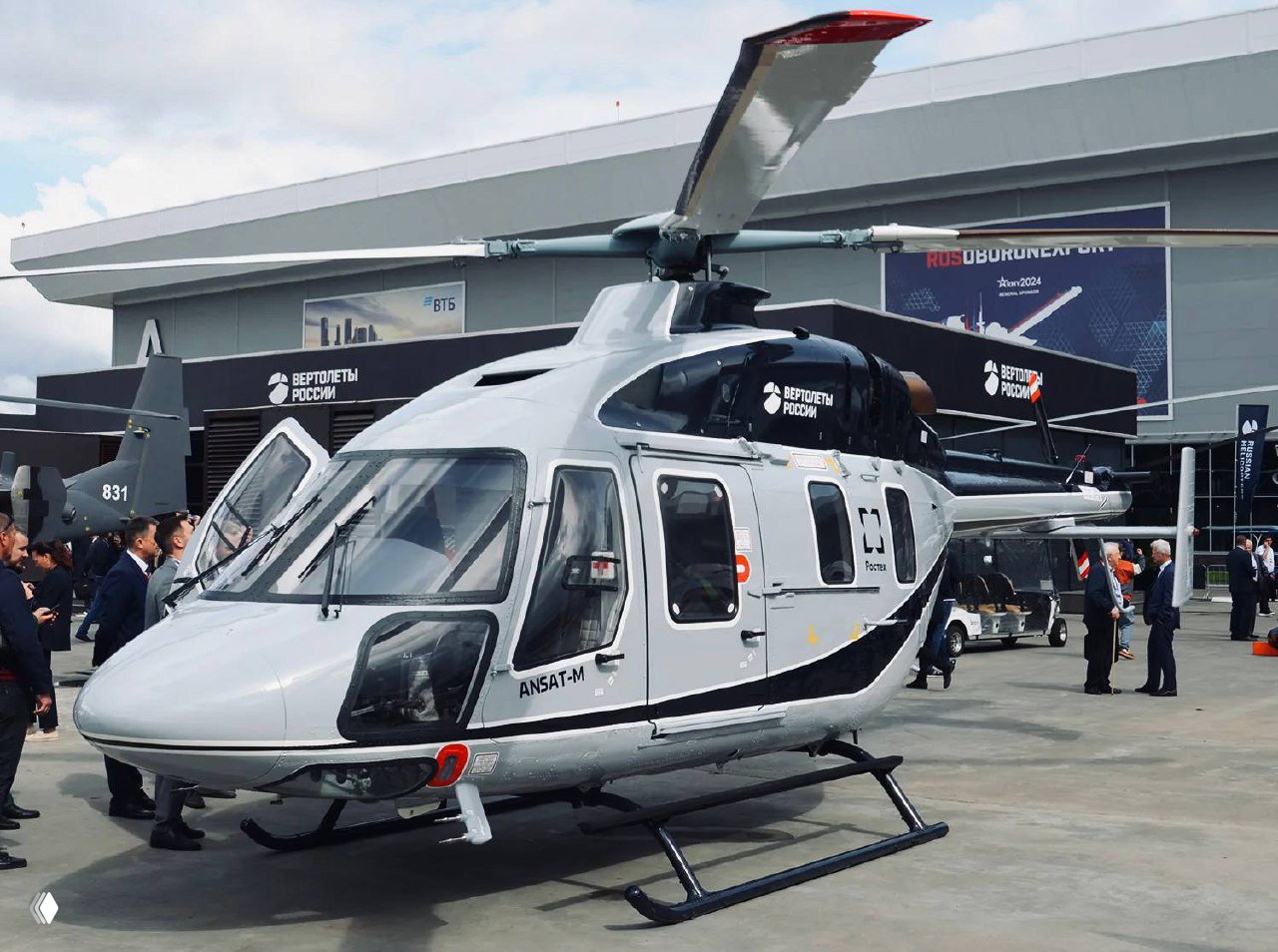 Вертолёт Ансат‑М на стоянке авиасалона с логотипами «Вертолёты России» и Ростеха, люди возле экспозиции и демонстрационные стенды.