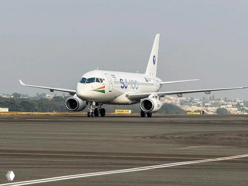 Superjet (SJ) на рулёжной дорожке в Хайдарабаде: вид спереди белого лайнера с ливреей и логотипом, самолёт на фоне аэродрома во время выставки.