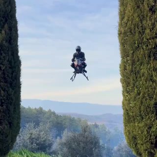 Фото: пилот на Volonaut Airbike зависает между высокими деревьями на фоне холмов, аппаратура и посадочные опоры чётко видны.
