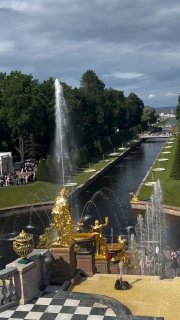 Вид на Петергоф: Большой каскад с золотыми статуями и фонтанами, пруд и аллеи парка под облачным небом.