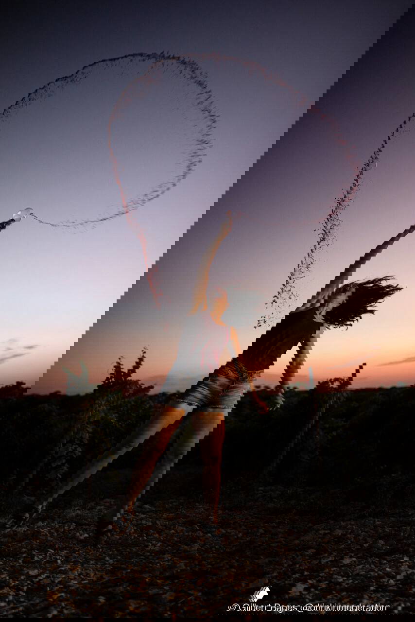 Женщина в винограднике подбрасывает бокал с вином; образуется эффектная дуга брызг на фоне закатного неба — динамичная фотография.