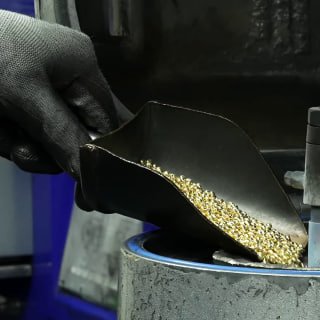 Gloved hand pouring a scoop of small gold granules into a jeweller's container in a workshop, showing raw precious-metal material handling.