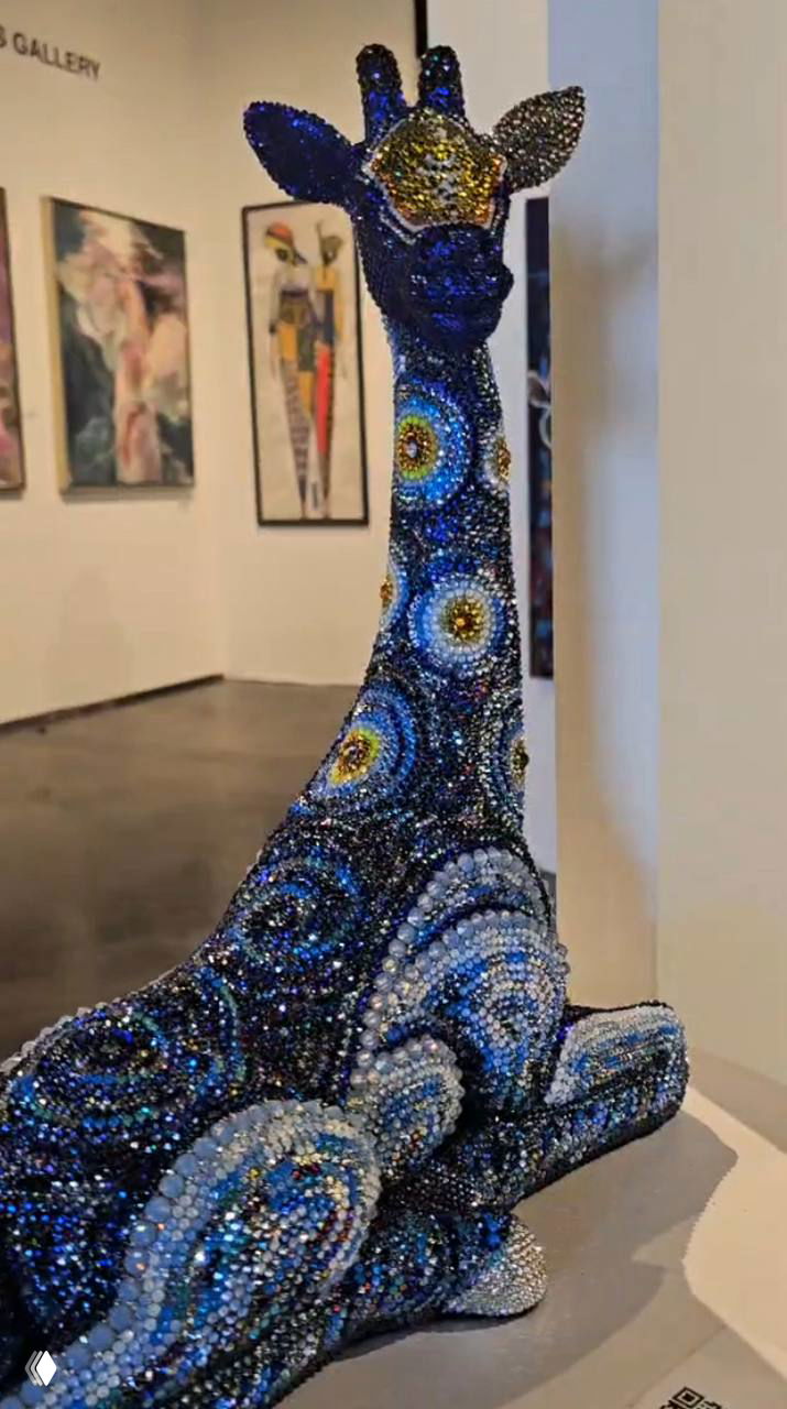 Статуэтка жирафа, полностью покрытая разноцветными кристаллами и узорной мозаикой, выставлена на пьедестале в интерьере галереи с картинами на стене.