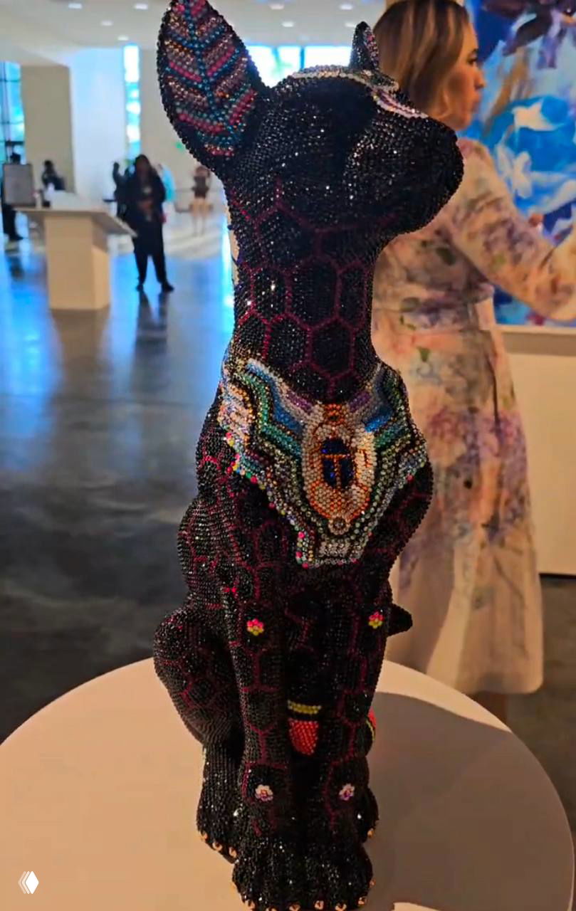 Обратная сторона тёмной скульптуры животного, вся в мелких стразах и цветных акцентах, на фоне посетителей галереи и светлого пространства.