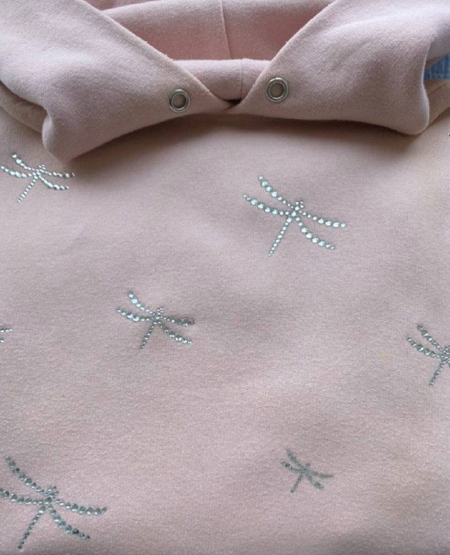 Крупный план светло-розовой толстовки с узором из мелких страз — похожие на стрекозы, видны люверсы капюшона.