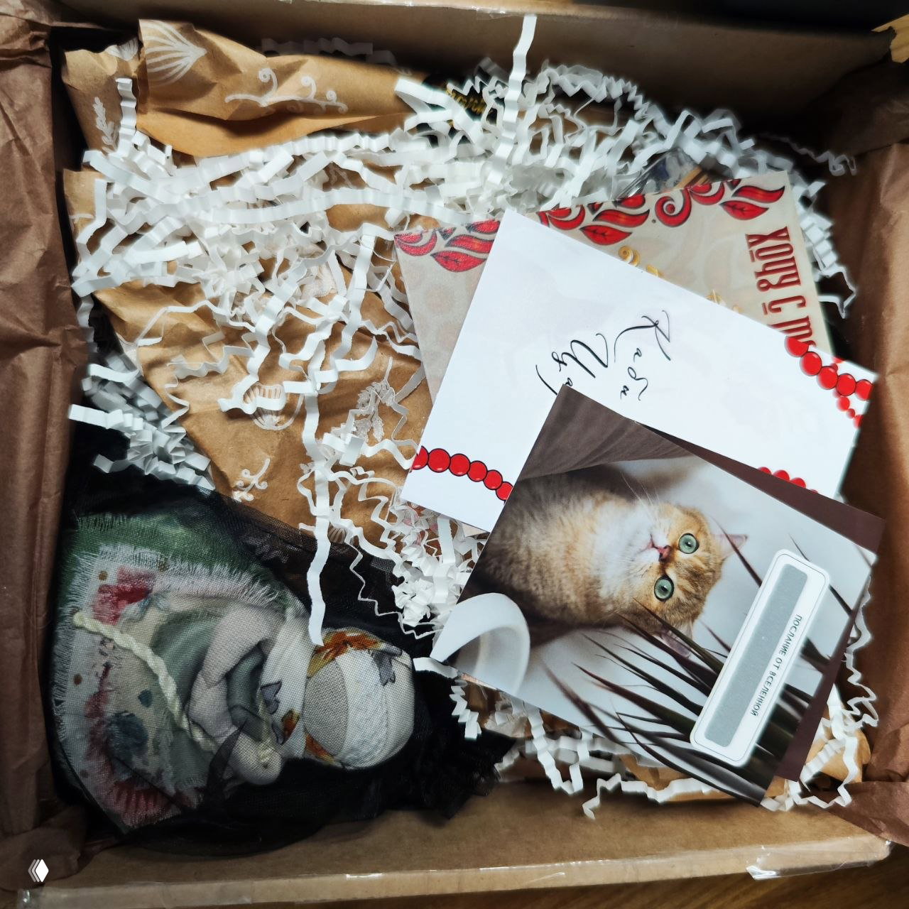 Вид сверху на короб с белым бумажным наполнителем, открытками и спрятанной обережной куклой — оформление и карточки внутри посылки.