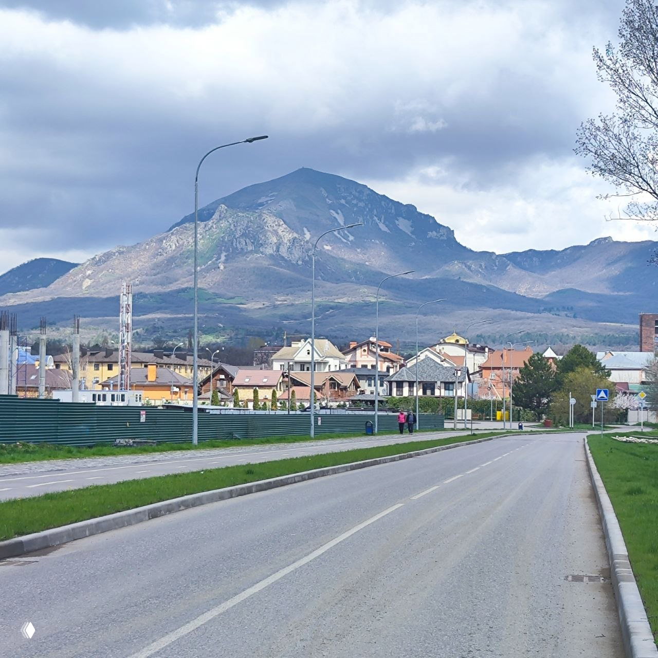 Панорамный вид на Машук и окрестности Пятигорска: пустая дорога и жилые дома на переднем плане, горы под тучным небом.