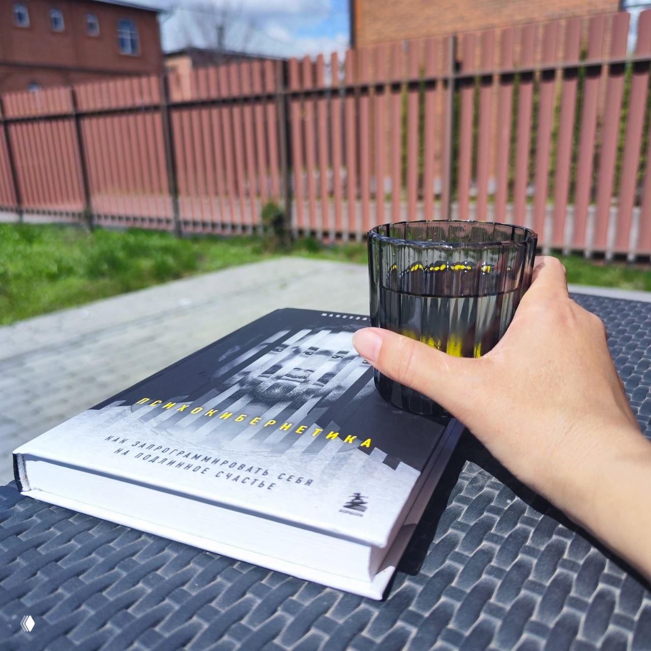 Книга и стакан чая на плетёном столе на террасе: крупный план руки, книга на русском языке и солнечное настроение весеннего отдыха.