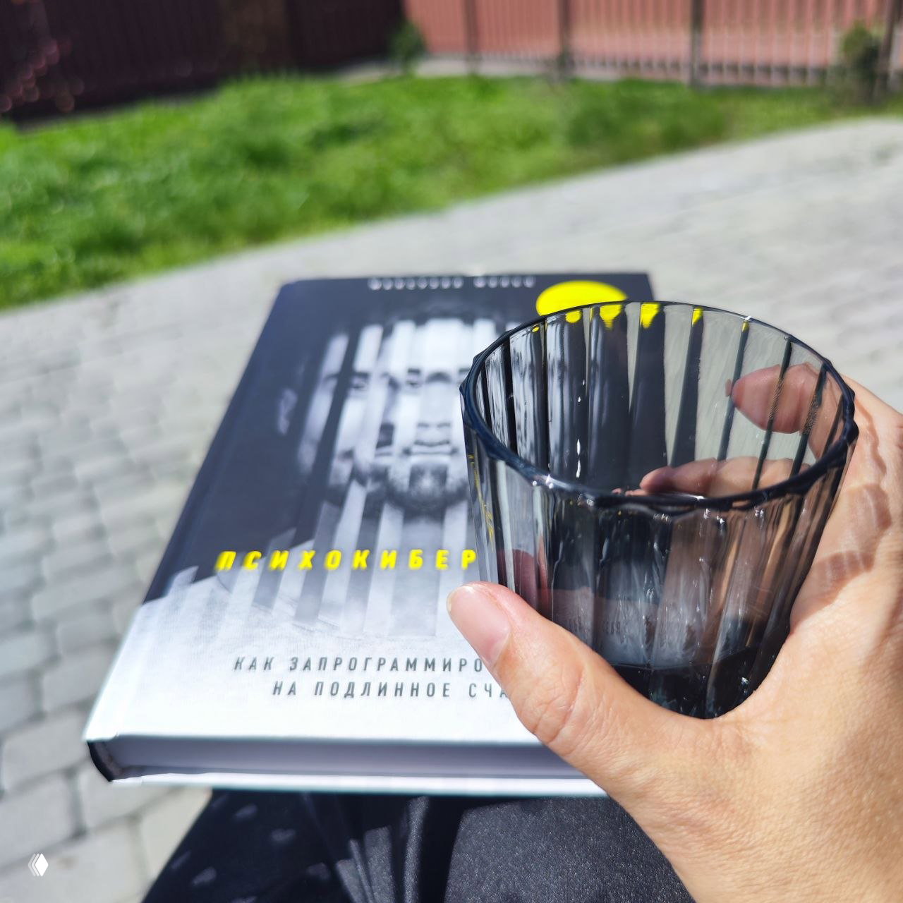 Стеклянный стакан в руке и книга на коленях на террасе: момент чтения и отдыха на свежем воздухе, размытый фон газона и плитки.