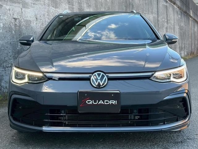 Вид спереди: Volkswagen Golf 2021 (гибрид), эмблема VW на капоте, светящиеся фары и решётка радиатора, машина на фоне бетонной стены, фронтальный ракурс.