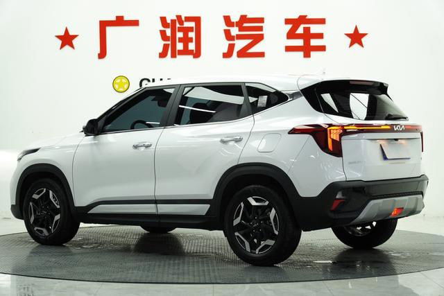 Kia Seltos белого цвета, вид сзади и в профиль на фоне стенда; заметны задние фонари, бампер и общий силуэт кроссовера в салоне.