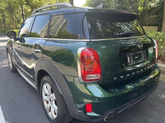 Задняя правая часть зелёного MINI Countryman 2019, вид сзади и кормы, фон — лиственный сквер и асфальт