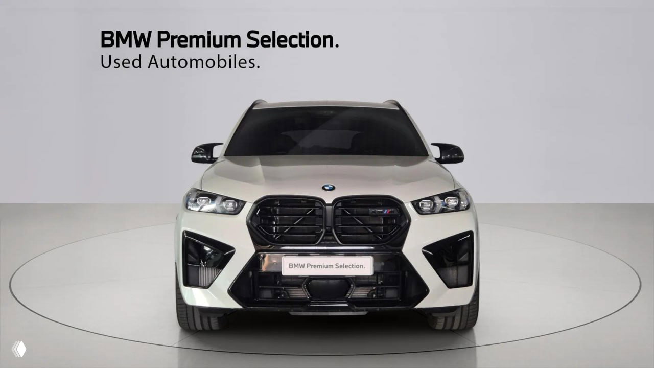 Фронтальный вид белого BMW X5 M Competition 2025: крупный кадр центральной решетки, фары и передний бампер в выставочном пространстве.