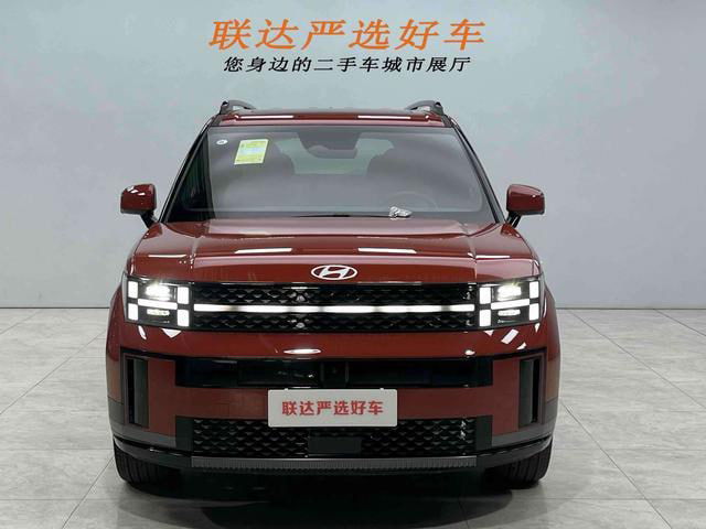 Hyundai Santa Fe 2024 крупный фронтальный план в выставочном помещении, включённые светодиодные фары и логотип на капоте видны чётко.