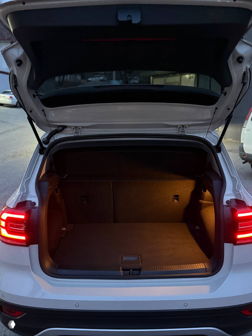 Задняя трёхчетвертная проекция VW T‑Cross с включёнными габаритными огнями, видны эмблема VW и модель на крышке багажника в городской обстановке.