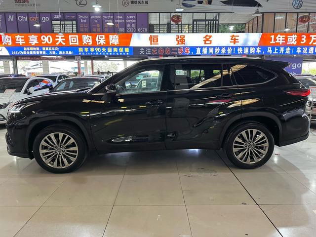 Боковой профиль чёрного Toyota Highlander 2022 в выставочном зале, видно диски, линию крыши и отражения рекламных баннеров над автомобилем.