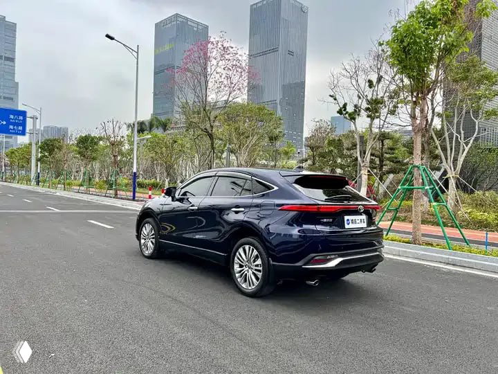 Тёмно-синий Toyota Harrier 2022 — общий задне-боковой ракурс, автомобиль припаркован на улице с современными зданиями и зелёными насаждениями.