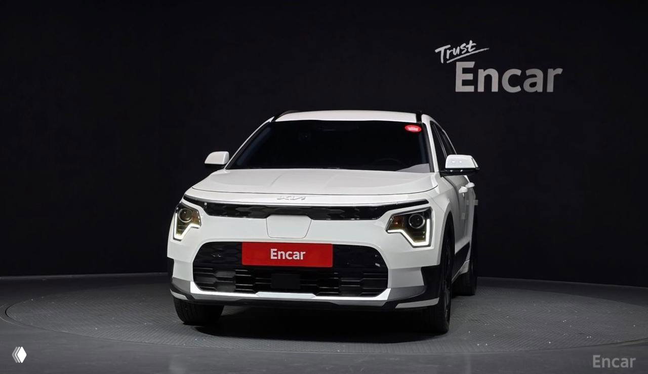 Передний вид белого Kia Niro в студии Encar: решётка радиатора, фары LED и красная табличка Encar на чёрном фоне, крупный план.