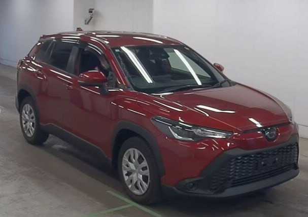 Toyota Corolla Cross красного цвета, ракурс спереди слева в помещении аукциона, видны передние фары, зеркало и боковая панель кузова.