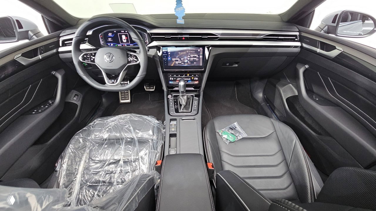 Интерьер Volkswagen Arteon 2022: руль с логотипом VW, цифровая приборная панель и центральный тачскрин, пластиковая защита на переднем сиденье.