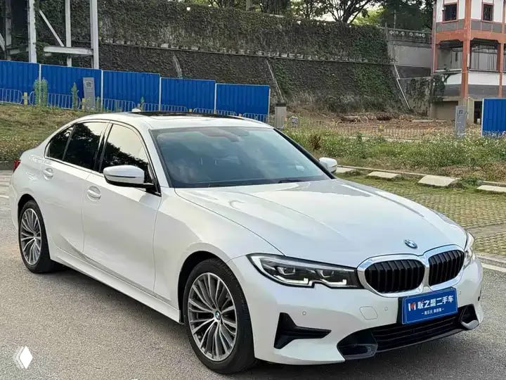 Белый BMW 3 Series 320i, боковой ракурс; автомобиль стоит на площадке с ограждением и зданиями на заднем плане.