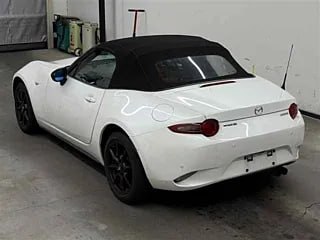 Белая Mazda Roadster вид сзади и в три четверти, видна задняя оптика, бампер и чёрный складывающийся верх, автомобиль на стоянке внутри помещения.