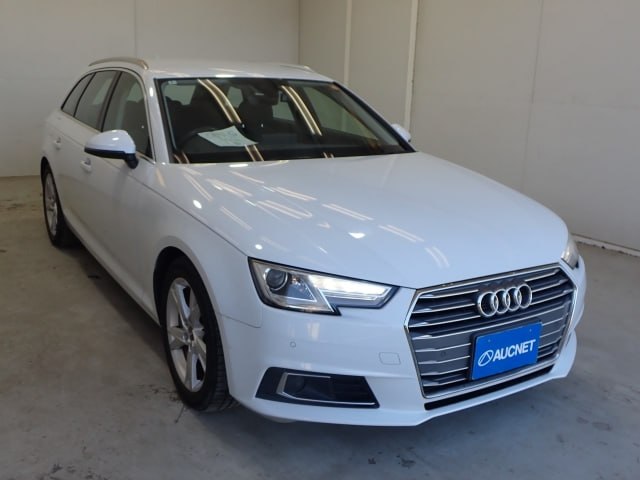 Передняя часть белого Audi A4 2018 в ангаре, крупный план на решетку радиатора и фары, автомобиль под естественным освещением.