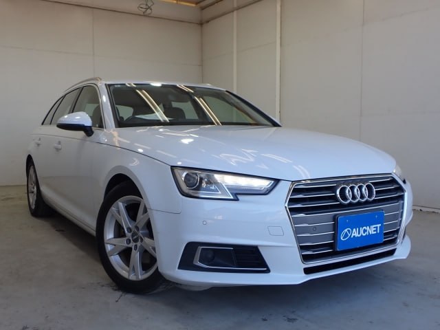 Белый Audi A4 2018 (универсал) в боковом ракурсе в светлом ангаре‑шоуруме; видно переднюю часть, фон и пластину с логотипом AUCNET.