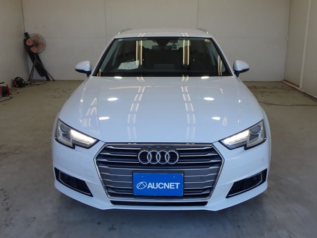 Фронтальный вид белого Audi A4 2018: ровная морда, включенные ДХО, видно отражения потолочных ламп в кузове.