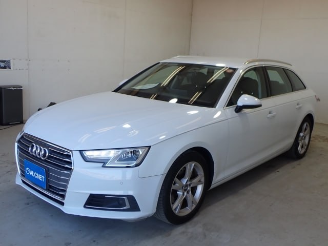 Трехчетвертной ракурс белого Audi A4 2018 спереди‑сбоку в ангаре; видны колеса, передний бампер и боковая линия кузова.