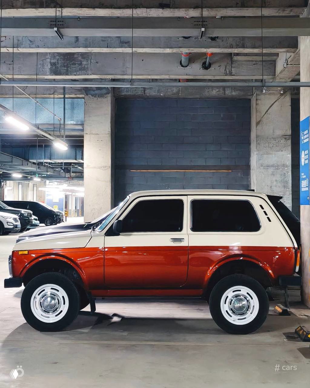 Боковой профиль двухцветной Нивы в паркинге — белая верхняя часть и тёплый коричневый низ, хромированные колпаки и ретро‑элементы оформления.